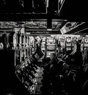 A dimly lit casino filled with rows of slot machines, some occupied by people. The machines' lights contrast with the dark surroundings, creating a vibrant yet subdued atmosphere.