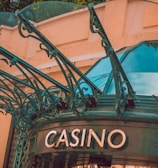 The image displays the entrance to a casino, characterized by detailed and ornate architectural elements with green metal structures and a sign that reads 'CASINO'. The background includes a glass dome and a peach-colored wall.
