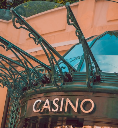 The image displays the entrance to a casino, characterized by detailed and ornate architectural elements with green metal structures and a sign that reads 'CASINO'. The background includes a glass dome and a peach-colored wall.
