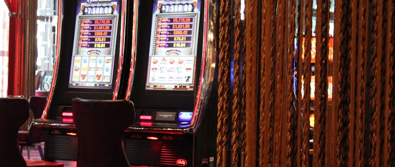 A pair of casino slot machines, each with detailed screens displaying various game outcomes and jackpot amounts. The chairs in front of the slot machines are dark and cushioned for comfort. The background features a hanging curtain made of golden ropes and other casino decorations are visible.