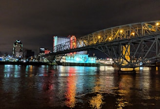 A brightly lit casino with neon signs and red lights is reflected in a river. A bridge stretches across the water, with an urban skyline visible in the background against an overcast night sky.