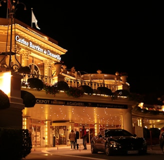 A luxurious casino at night, illuminated with warm, golden lights. The building features elegant architecture with columns and decorative elements. A few people are walking towards the entrance, and there are a couple of parked cars nearby.