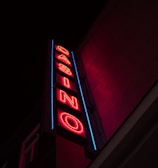 A neon sign is prominently displayed on the side of a building. The sign is vertical and spells out the word 'CASINO' in bold, red neon letters. Surrounding the letters is a blue outline that adds to the vibrancy of the sign against the otherwise dark facade of the building.