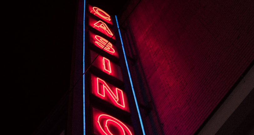 A neon sign is prominently displayed on the side of a building. The sign is vertical and spells out the word 'CASINO' in bold, red neon letters. Surrounding the letters is a blue outline that adds to the vibrancy of the sign against the otherwise dark facade of the building.