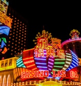 Bright neon lights and colorful signage adorn the exterior of Casino Lisboa, creating a vibrant and lively atmosphere. The façade features bold, multicolored designs with a mix of English and Chinese characters prominently displayed. The lights form intricate patterns and shapes, contributing to the energetic ambiance of the scene.