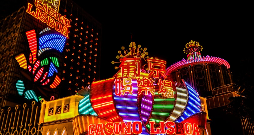 Bright neon lights and colorful signage adorn the exterior of Casino Lisboa, creating a vibrant and lively atmosphere. The façade features bold, multicolored designs with a mix of English and Chinese characters prominently displayed. The lights form intricate patterns and shapes, contributing to the energetic ambiance of the scene.