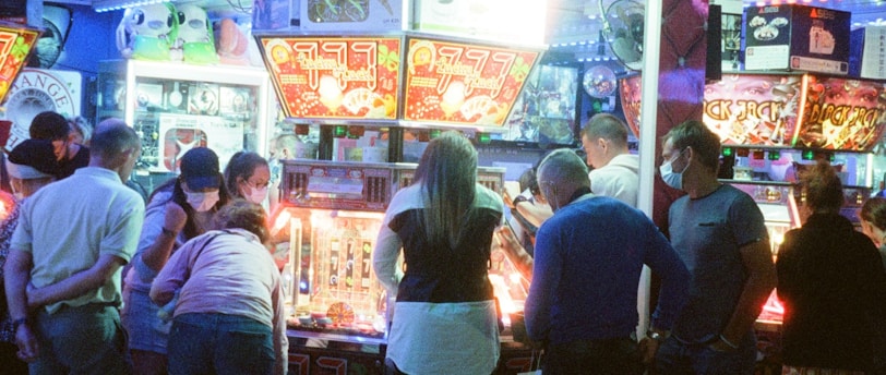 A group of people standing in front of an illuminated amusement arcade, engaging with various gaming machines. The environment is bustling with activity, and colorful lights create a vibrant atmosphere. The ceiling is decorated with large, playful images of dice and other game-related graphics, enhancing the lively mood.