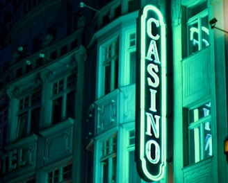 A vibrant neon sign displaying the word 'CASINO' is prominently illuminated against the facade of a building, showcasing intricate architectural details and a classic design. The sign emits a bright teal glow, casting light on the surrounding area and creating a lively atmosphere at night.