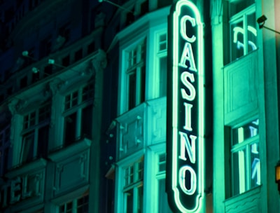A vibrant neon sign displaying the word 'CASINO' is prominently illuminated against the facade of a building, showcasing intricate architectural details and a classic design. The sign emits a bright teal glow, casting light on the surrounding area and creating a lively atmosphere at night.