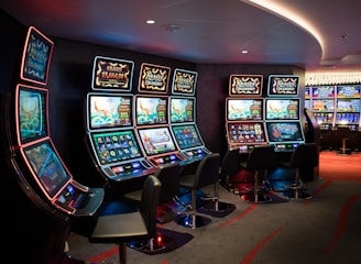 A row of brightly lit slot machines in a casino setting, each with colorful displays and various game themes. The machines are lined up against a dark wall and are unoccupied, with cushioned chairs placed in front of them. The area is dimly lit except for the glow from the screens, creating a vibrant and engaging atmosphere.