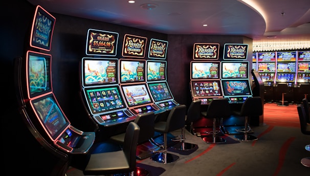 A row of brightly lit slot machines in a casino setting, each with colorful displays and various game themes. The machines are lined up against a dark wall and are unoccupied, with cushioned chairs placed in front of them. The area is dimly lit except for the glow from the screens, creating a vibrant and engaging atmosphere.