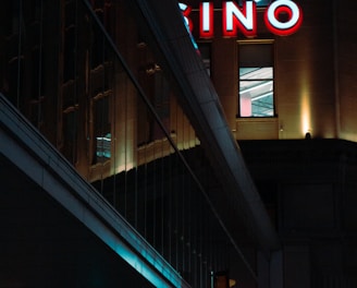 A night scene featuring a building with illuminated neon signage displaying 'CASINO'. The environment is urban with reflections on glass windows and glowing street traffic lights below. The atmosphere is dimly lit, with artificial lighting casting vibrant colors.
