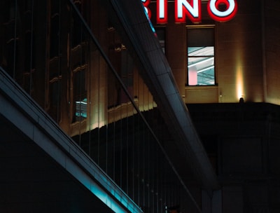A night scene featuring a building with illuminated neon signage displaying 'CASINO'. The environment is urban with reflections on glass windows and glowing street traffic lights below. The atmosphere is dimly lit, with artificial lighting casting vibrant colors.