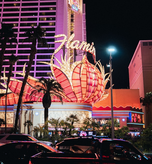 A brightly lit casino with neon lights in vibrant red, pink, and blue, featuring a large flamingo signage. Palm trees are visible in the foreground with vehicles passing by on the street at night.