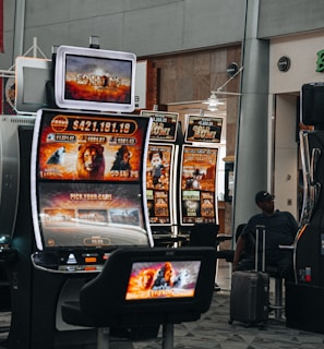 A collection of slot machines is situated inside a building with bright, colorful screens displaying various games and gaming themes. A person is seated nearby, seemingly relaxed with luggage beside them, and the atmosphere is lively with electronic displays.