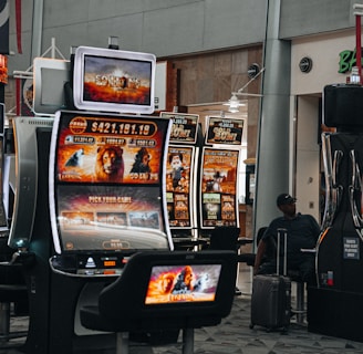 A collection of slot machines is situated inside a building with bright, colorful screens displaying various games and gaming themes. A person is seated nearby, seemingly relaxed with luggage beside them, and the atmosphere is lively with electronic displays.